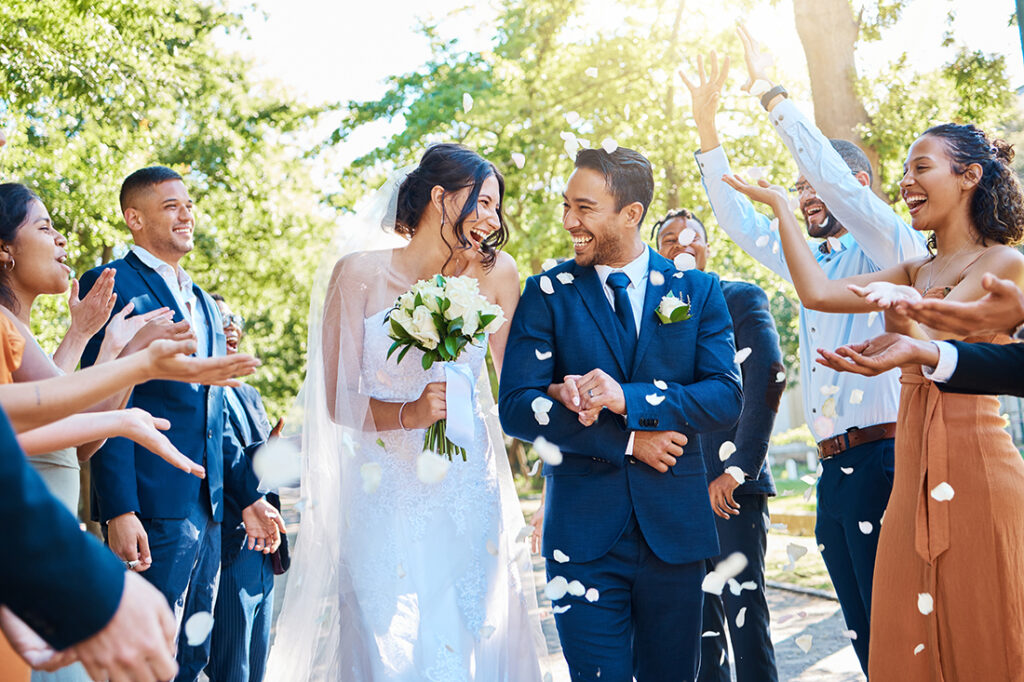 Happy, wedding ceremony and couple walking with petals and guests throw in celebration of romance. Romantic, flowers and bride with bouquet and groom with crowd of friends, family, and co-workers celebrating at outdoor marriage event.