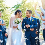 Happy, wedding ceremony and couple walking with petals and guests throw in celebration of romance. Romantic, flowers and bride with bouquet and groom with crowd of friends, family, and co-workers celebrating at outdoor marriage event.