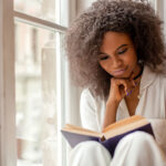 african american girl with reading a book sitting on the windowsill